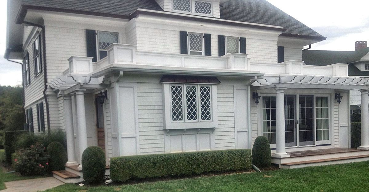 White shingle home with pergolas, round columns and a diamond-paned bay window